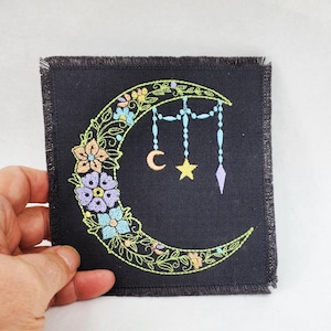 May include: Black square patch with a crescent moon design. The moon is adorned with colorful embroidered flowers and hanging beads, a crescent moon, a star, and a diamond shape. The patch has a frayed edge.