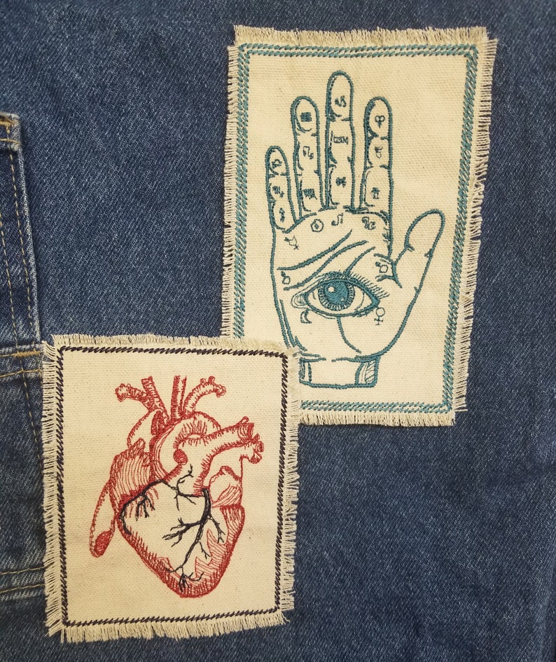 Puede incluir: Dos parches bordados sobre un fondo de mezclilla azul. Un parche presenta un coraz&oacute;n anat&oacute;mico rojo, el otro una mano con un ojo en la palma, con bordado turquesa.