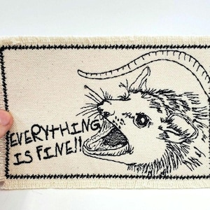 May include: A white canvas patch with black embroidery of an opossum with its mouth open and the text "EVERYTHING IS FINE!"