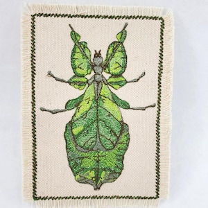 May include: Embroidered patch featuring a detailed green leaf insect design. The insect is depicted with intricate leaf-like wings and body, in shades of green and brown. The patch is bordered with a dark green stitched edge and has a cream-colored background.