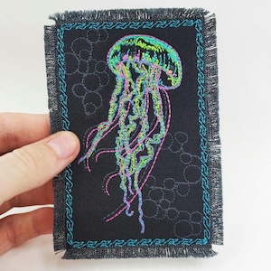 Puede incluir: Un parche de tela negro con borde de flecos presenta una medusa bordada con tentáculos de color verde neón, rosa y azul.