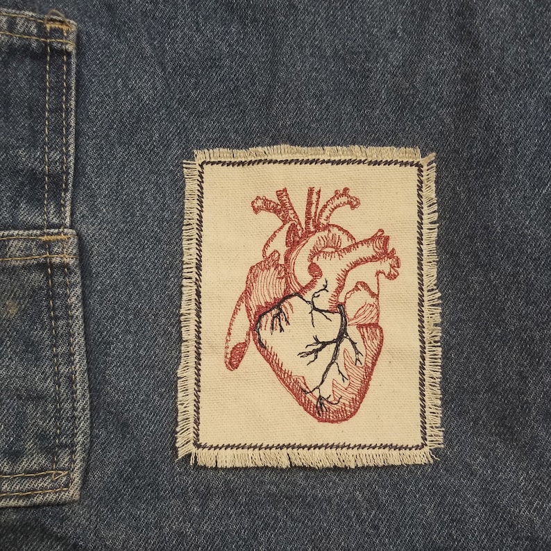 Puede incluir: Un coraz&oacute;n anat&oacute;mico rojo bordado en un parche de tela beige con bordes deshilachados. El parche est&aacute; cosido sobre un fondo de mezclilla azul.
