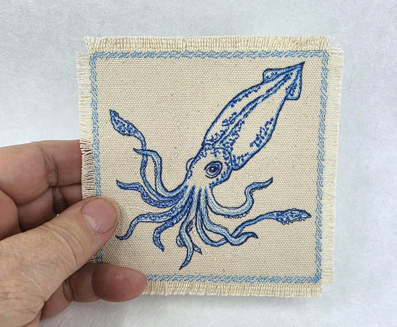 May include: A square fabric patch featuring a blue embroidered squid design on a natural beige canvas background. The patch has a frayed edge and a decorative blue stitched border with a repeating wave pattern surrounding the central squid.