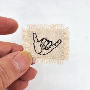 May include: A small, rectangular, off-white fabric patch with frayed edges. The patch features a black embroidered outline of a hand making the shaka sign, with the thumb and pinky finger extended while the middle three fingers are curled inward.