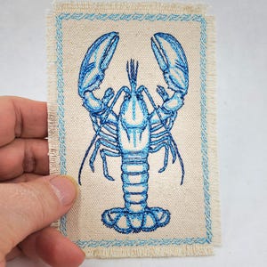 May include: Embroidered patch featuring a blue lobster design on a beige fabric. The lobster is intricately detailed with a light blue outline and shading. The patch has a frayed edge and a light blue stitched border. The patch is rectangular.