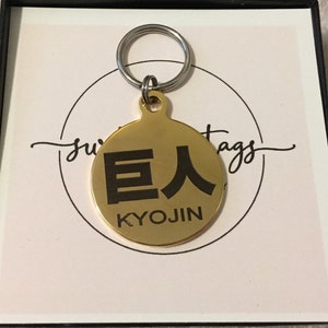 May include: A gold-colored metal dog tag with a black engraved design. The tag features the Japanese characters "巨人" (Kyojin) and the word "KYOJIN" in English.