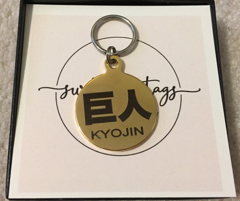 May include: A gold-colored metal dog tag with a black engraved design. The tag features the Japanese characters "巨人" (Kyojin) and the word "KYOJIN" in English.