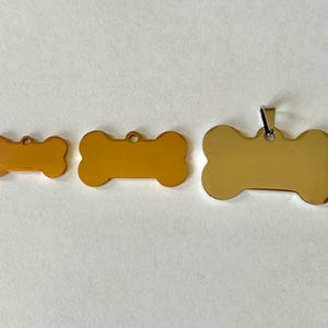 May include: Three gold bone-shaped dog tags of different sizes. The largest tag has a silver ring for attaching to a collar.