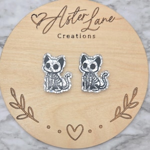 May include: A pair of stud earrings shaped like sitting cats with a white skeleton print on a black background. The cats have large black eyes and a glittery finish. The earrings are displayed on a round wooden card with the text Aster Lane Creations.