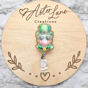 May include: A green leprechaun with a gold glitter hat, sunglasses, and a bubblegum bubble, on a white glitter badge reel with a clear plastic strap and a white glitter charm. The badge reel is attached to a wooden circle with the text "Aster Lane Creations" and a heart.