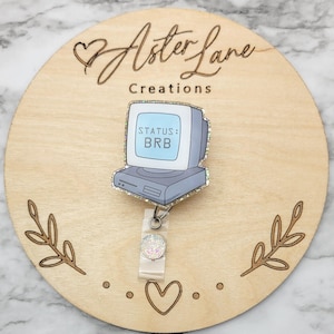 May include: A light blue and gray computer screen with the text "STATUS: BRB" on a white background. The computer is attached to a clear plastic badge holder with a sparkly iridescent charm.