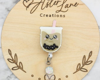 May include: A brown and white boba tea charm with a pink straw and a cute face. The charm is attached to a clear plastic badge holder with a sparkly white button. The charm is on a wooden background with the text "Aster Lane Creations".