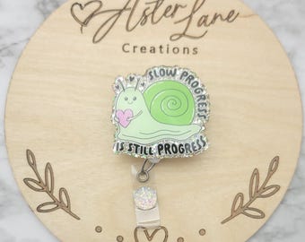Può includere: Un porta badge a forma di lumaca verde chiaro con la scritta "SLOW PROGRESS IS STILL PROGRESS". La lumaca tiene in mano un cuore rosa. Il porta badge ha un accento glitterato ed è attaccato a una base rotonda di legno con la scritta "Aster Lane Creations".