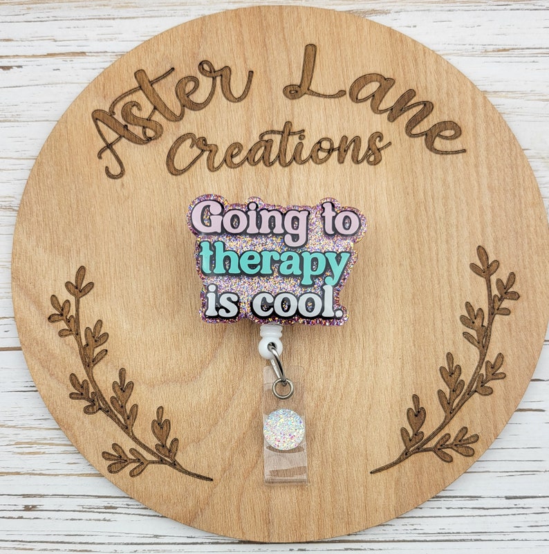 Op de afbeelding: Een glitterende badgehouder in roze, turquoise en wit met de tekst "Therapie is cool."