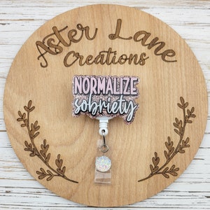May include: A pink and white glitter badge holder with a white retractable reel and a clear badge holder. The badge holder has the text "NORMALIZE sobriety" in white lettering.