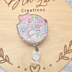 May include: A circular badge reel featuring a glittery finish and a colorful graphic with the text "in my SPEECH THERAPY era". The design includes a pink bow, a clipboard, a magnifying glass, headphones, and a set of teeth, all in pastel tones.