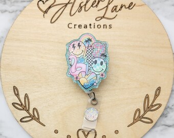 May include: A teal blue glitter badge reel with a colorful design featuring a flamingo, smiley faces, palm trees, and a rainbow. The text "Aster Lane Creations" is on the wooden background.