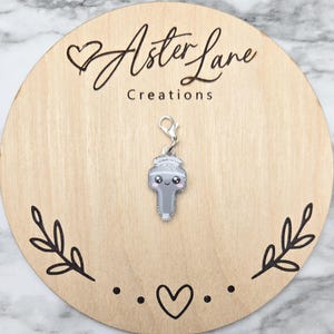 May include: A silver-colored charm with a cute, smiling face and a crown design. The charm is attached to a silver clasp. The charm is displayed on a wooden round board with the text "Aster Lane Creations".