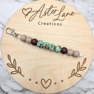 May include: A wooden circle with the text "Aster Lane Creations" and a heart design. A beaded keychain with brown and green beads is displayed on the circle.