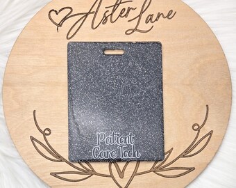 May include: A round wooden plaque with the text "Aster Lane" engraved on it. A black and white speckled name tag with the text "Patient Care Tech" is attached to the plaque.