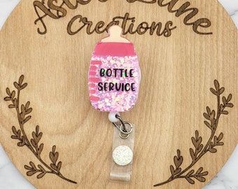 May include: Pink glitter baby bottle badge holder with the text "Bottle Service" on a wooden background.