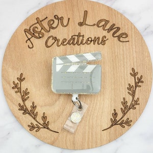 May include: A gray and white glitter film clapboard badge reel with a clear plastic badge holder and a silver metal retractable badge clip. The badge reel is on a wooden background with the text "Aster Lane Creations".