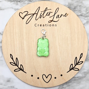 May include: Green gummy bear charm with a silver clip. The charm is on a wooden background with the text "Aster Lane Creations".