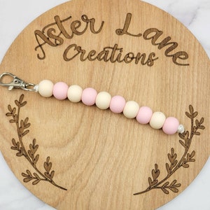 May include: A pink and cream silicone beaded keychain with a silver clip. The keychain is on a wooden background with the text "Aster Lane Creations" engraved on it.