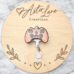 May include: A badge reel featuring a peach-colored pumpkin shaped like a video game controller with black button details and a dark glittery border. The badge reel has a white plastic retractable cord and a clear strap with a small white glittery accent.