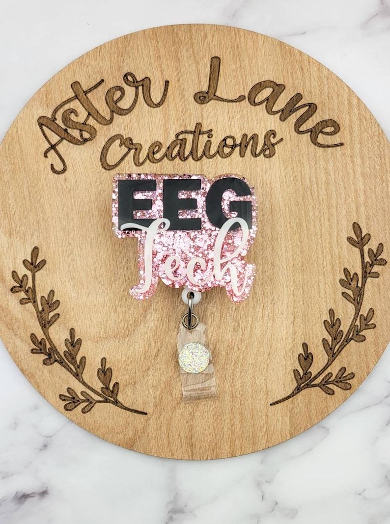 May include: Pink glitter badge reel with black text that reads "EEG Tech" and a clear retractable badge holder with a sparkly charm. The badge reel is on a wooden background with the words "Aster Lane Creations" engraved on it.