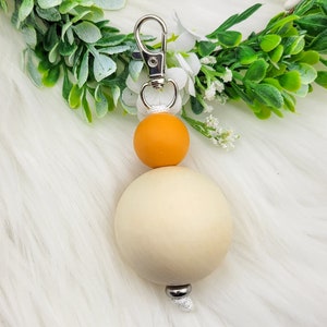 May include: A beige and orange silicone beaded keychain with a silver metal clasp. The keychain is hanging from a white fuzzy surface.