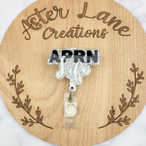 May include: A white glitter badge reel with a black and white "APRN Life" charm. The reel is on a wooden background with the words "Aster Lane Creations" engraved on it.