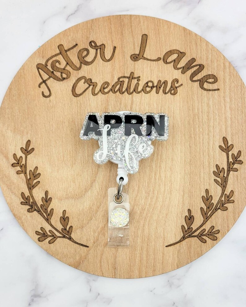 May include: A white glitter badge reel with a black and white "APRN Life" charm. The reel is on a wooden background with the words "Aster Lane Creations" engraved on it.