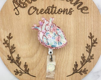May include: A pink glitter heart with a floral pattern, attached to a white retractable badge reel. The reel has a clear plastic cover with a glittery circle inside. The heart is on a wooden background with the text "Aster Lane Creations" engraved on it.
