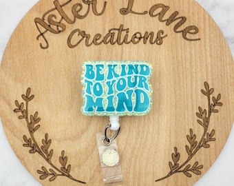 May include: A teal blue glitter badge reel with the text "BE KIND TO YOUR MIND" in white. The reel is attached to a clear plastic badge holder with a sparkly charm.