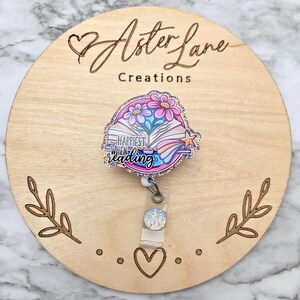May include: A pink and blue glitter badge reel with a book, flowers, and a shooting star. The text on the reel says "Happiest When Reading".