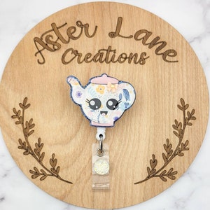 May include: A white teapot with a floral pattern and a pink lid. The teapot has a cute cartoon face with big eyes and a smile. The teapot is attached to a clear plastic badge holder with a sparkly glitter charm.