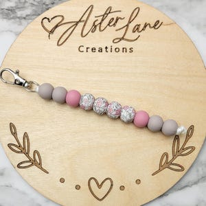 May include: A gray and pink beaded keychain with a floral pattern. The keychain has a silver key ring and is attached to a wooden circle with the text "Aster Lane Creations".