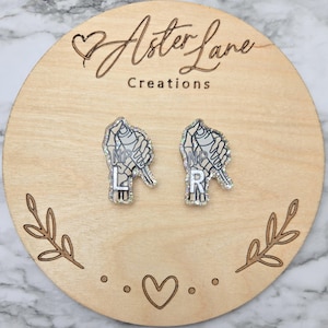 May include: A pair of acrylic stud earrings featuring a skeletal hand holding a spray paint can. The left earring displays the letters NP and L, while the right earring displays the letters NP and R. The design has a silver glitter finish.
