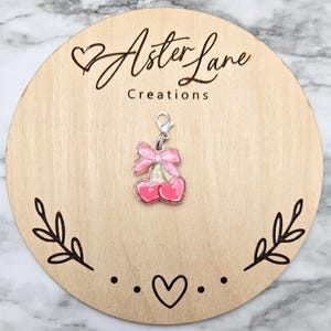 May include: A pink cherry charm with a silver clasp and a pink bow. The charm is on a wooden round with the text "Aster Lane Creations". The wooden round has a heart and leaf design.