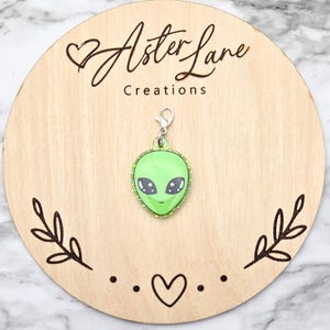 May include: Green alien charm with glitter, featuring a smiling face with closed eyes and white stars. The charm is attached to a silver chain and is displayed on a wooden background with the text "Aster Lane Creations".