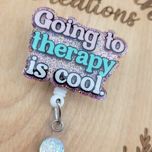 Op de afbeelding: Een roze en blauwe glitter badgehouder met de tekst "Going to therapy is cool." De badgehouder heeft een witte intrekbare rol en een transparante plastic hoes met een glinsterende regenboogkleurige cirkel.