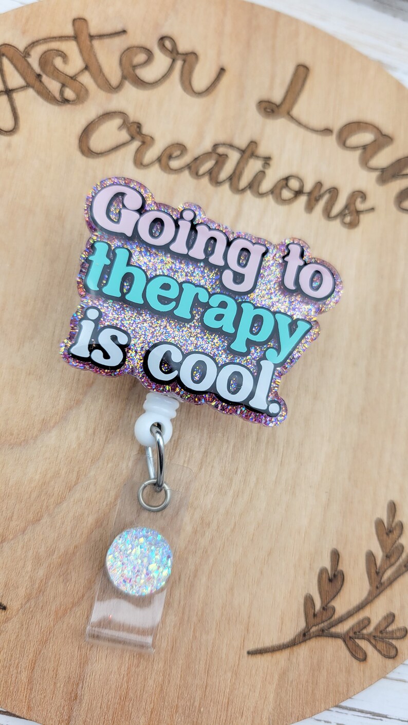 Op de afbeelding: Een roze en blauwe glitter badgehouder met de tekst "Going to therapy is cool." De badgehouder heeft een witte intrekbare rol en een transparante plastic hoes met een glinsterende regenboogkleurige cirkel.