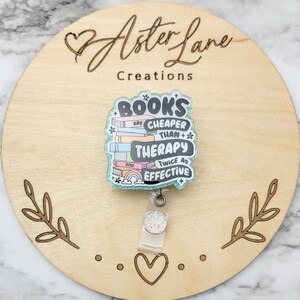 May include: A teal blue glitter badge holder with a stack of colorful books and the text "Books are cheaper than therapy and twice as effective".
