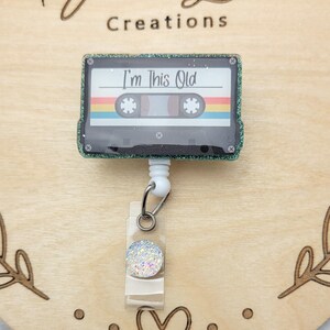 May include: A green glitter cassette tape with a white background and the text "I'm This Old" on it. The cassette tape is attached to a white retractable badge holder with a clear plastic cover and a sparkly white button.