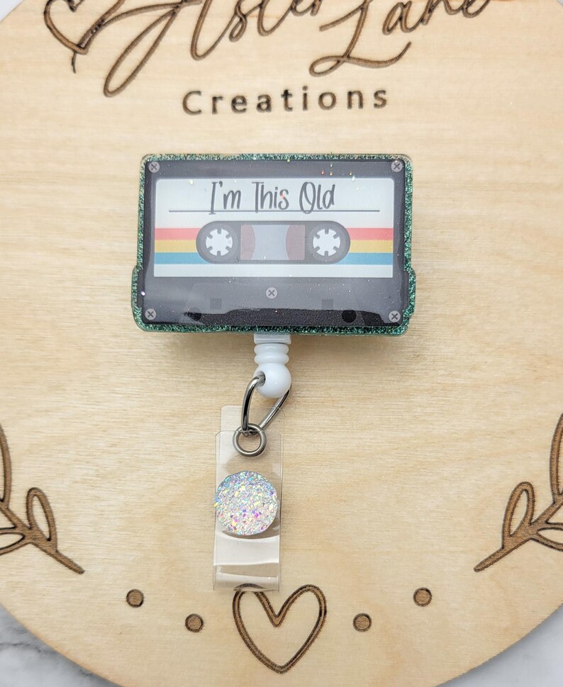 May include: A green glitter cassette tape with a white background and the text "I'm This Old" on it. The cassette tape is attached to a white retractable badge holder with a clear plastic cover and a sparkly white button.