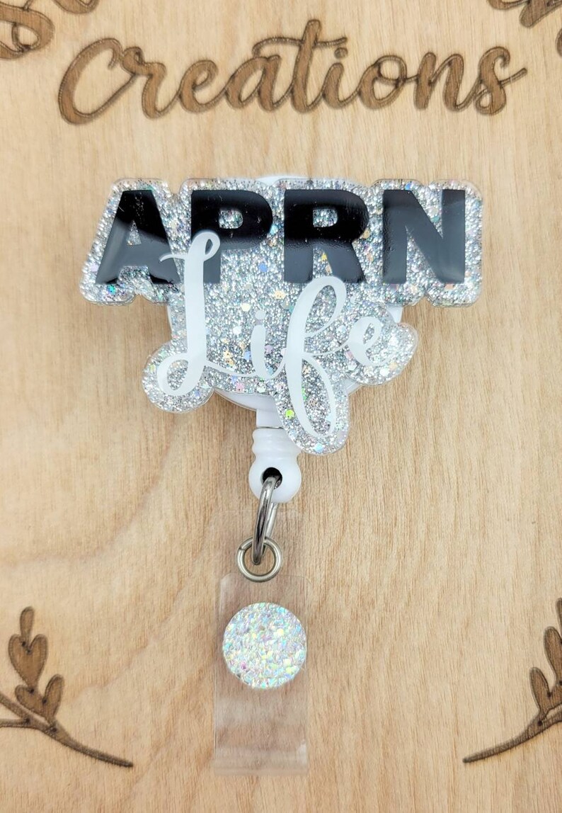 May include: A silver glitter badge reel with a black and white 'APRN Life' design. The reel has a clear plastic badge holder with a silver glitter circle.