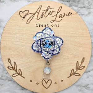 May include: A sparkly blue and silver atom charm with a cute cartoon face, attached to a clear retractable badge holder. The charm is on a wooden background with the text "Aster Lane Creations".