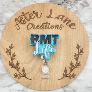 May include: A teal blue and black glitter badge reel with the text "EMT Life". The reel is attached to a wooden circle with the text "Aster Lane Creations" engraved on it.