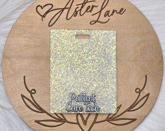 May include: A white wooden circle with the text "Aster Lane" engraved on it. A heart shape is above the text. A white glitter card with the text "Patient Care Tech" is in the center of the circle.